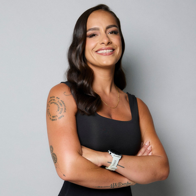 Smiling person with dark wavy hair, black tank top, and visible tattoos, arms crossed against a gray background