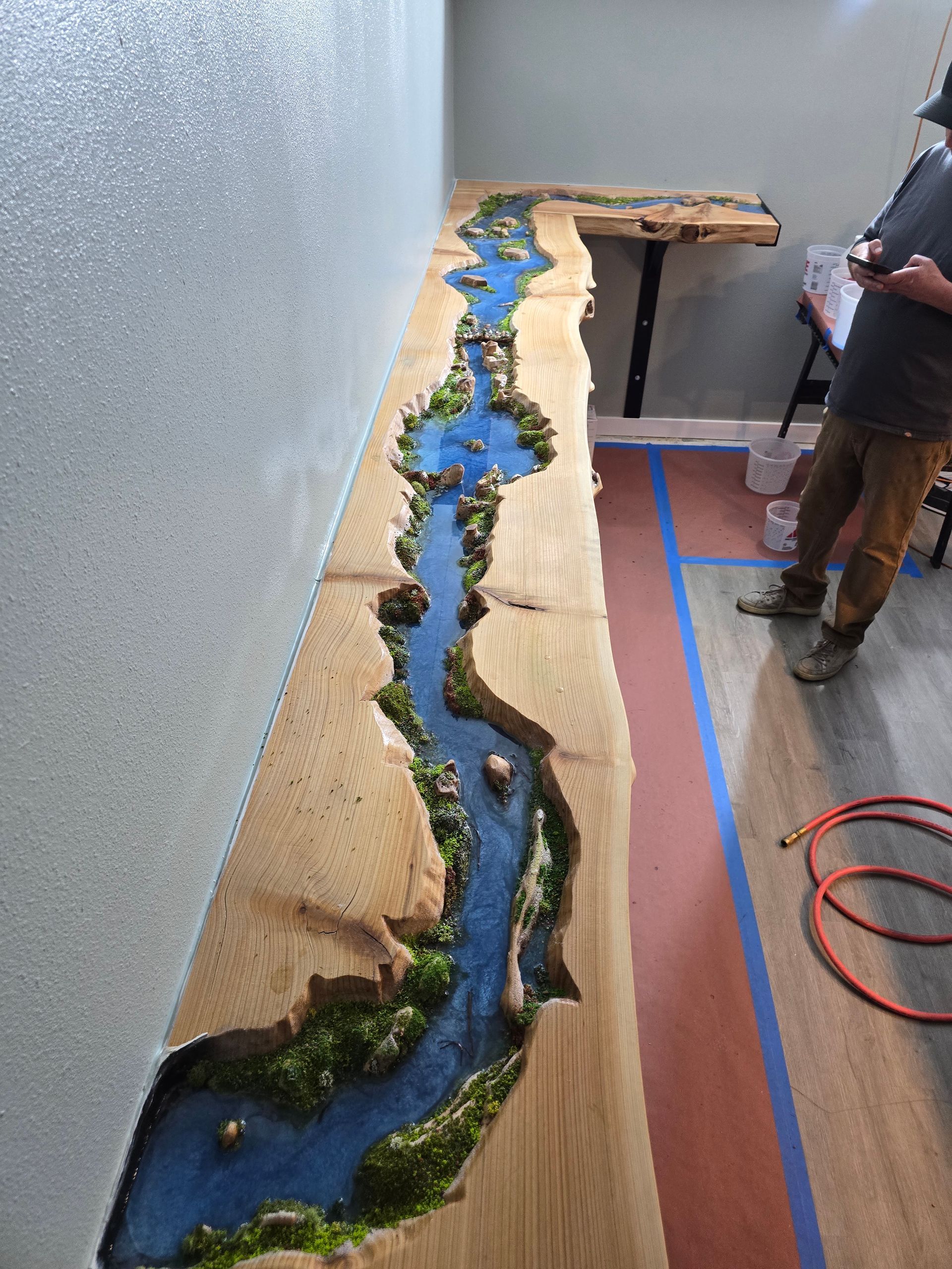 Long narrow model river landscape with blue water, green banks, and a person standing beside it in a room