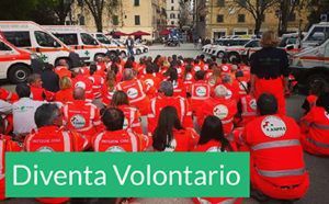 Volontari con giubbotti rossi siedono in fila all'aperto accanto alle ambulanze; uno striscione recita "Diventa Volontaria".