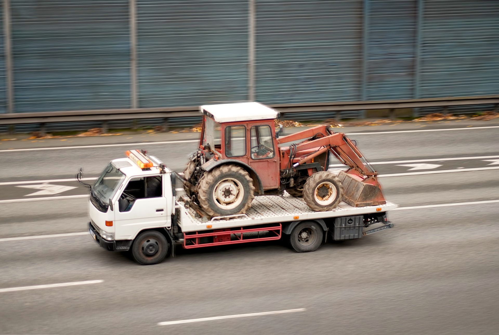 A white tow truck hauling a red tractor by a highway sound wall. A white tow truck hauling a red tractor by a highway sound wall.