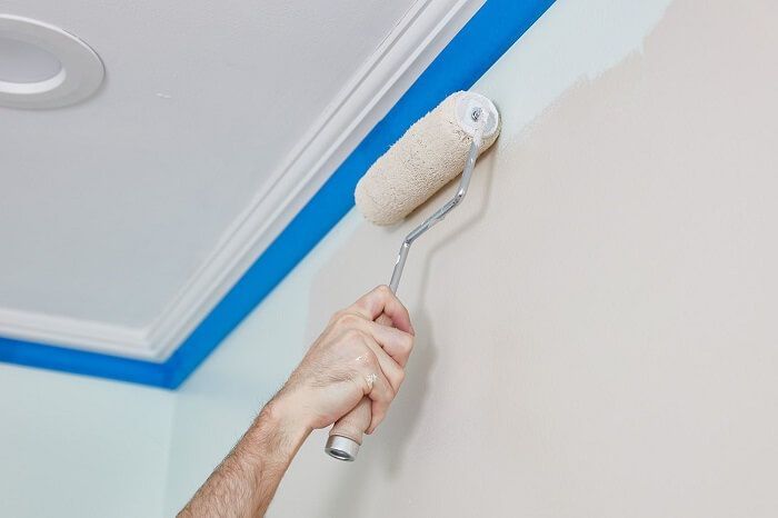 A person painting a wall with a roller. Blue painter's tape borders the ceiling trim.