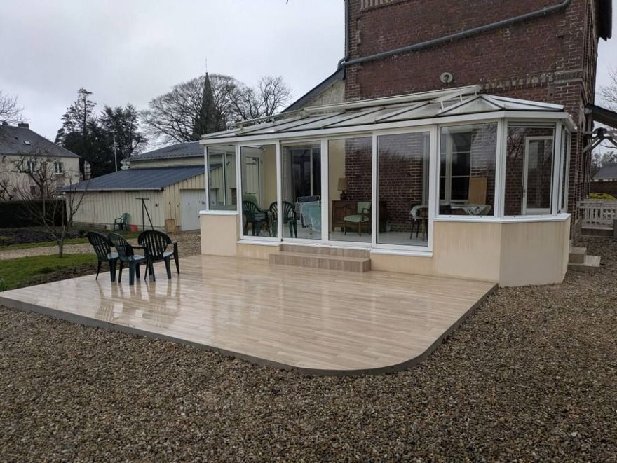 Une maison avec une véranda vitrée et une terrasse, entourée de gravier. Journée nuageuse.