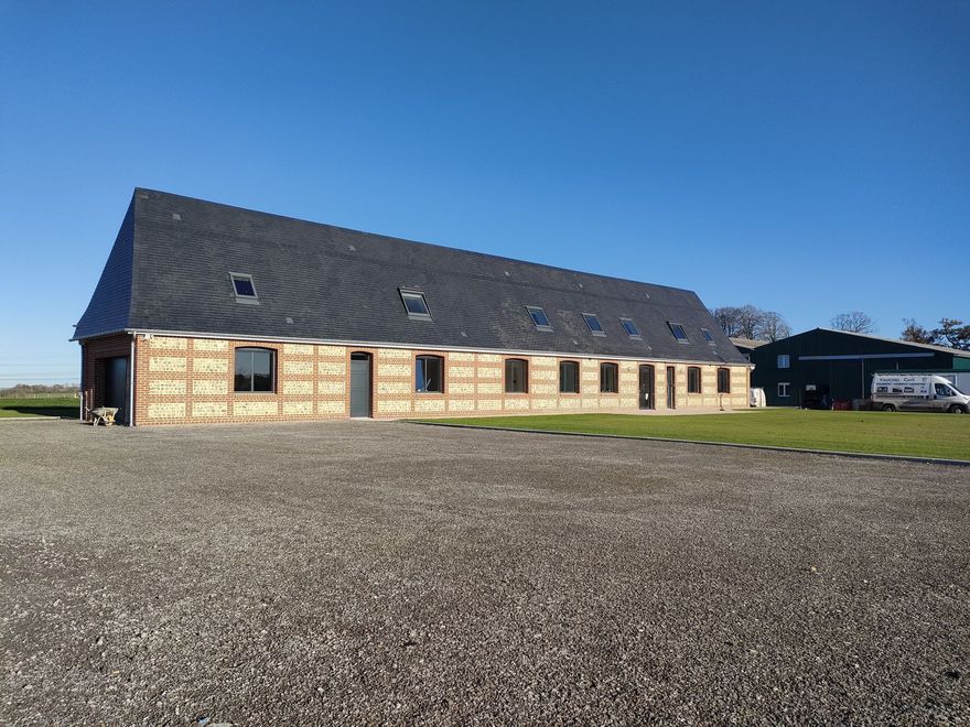 Bâtiment long, brun clair, avec toit, fenêtres et portes sombres, sur un sol gravillonné, sous un ciel bleu.