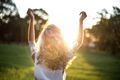 Femme, les bras levés, dos à la caméra, illuminée par la vive lumière du soleil dans un champ herbeux.