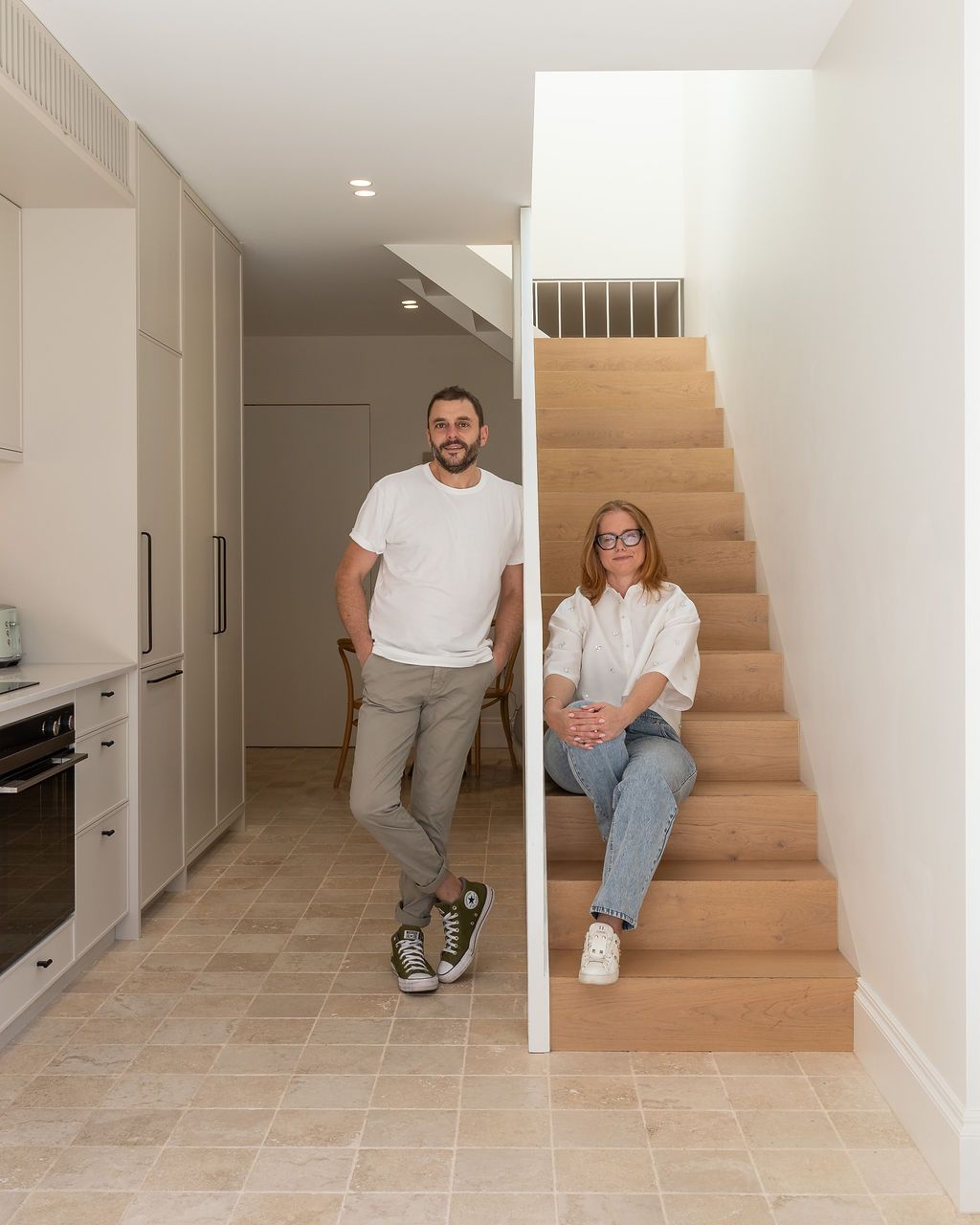 Two people posing in a bright hallway beside a staircase, one standing and one sitting on the steps.