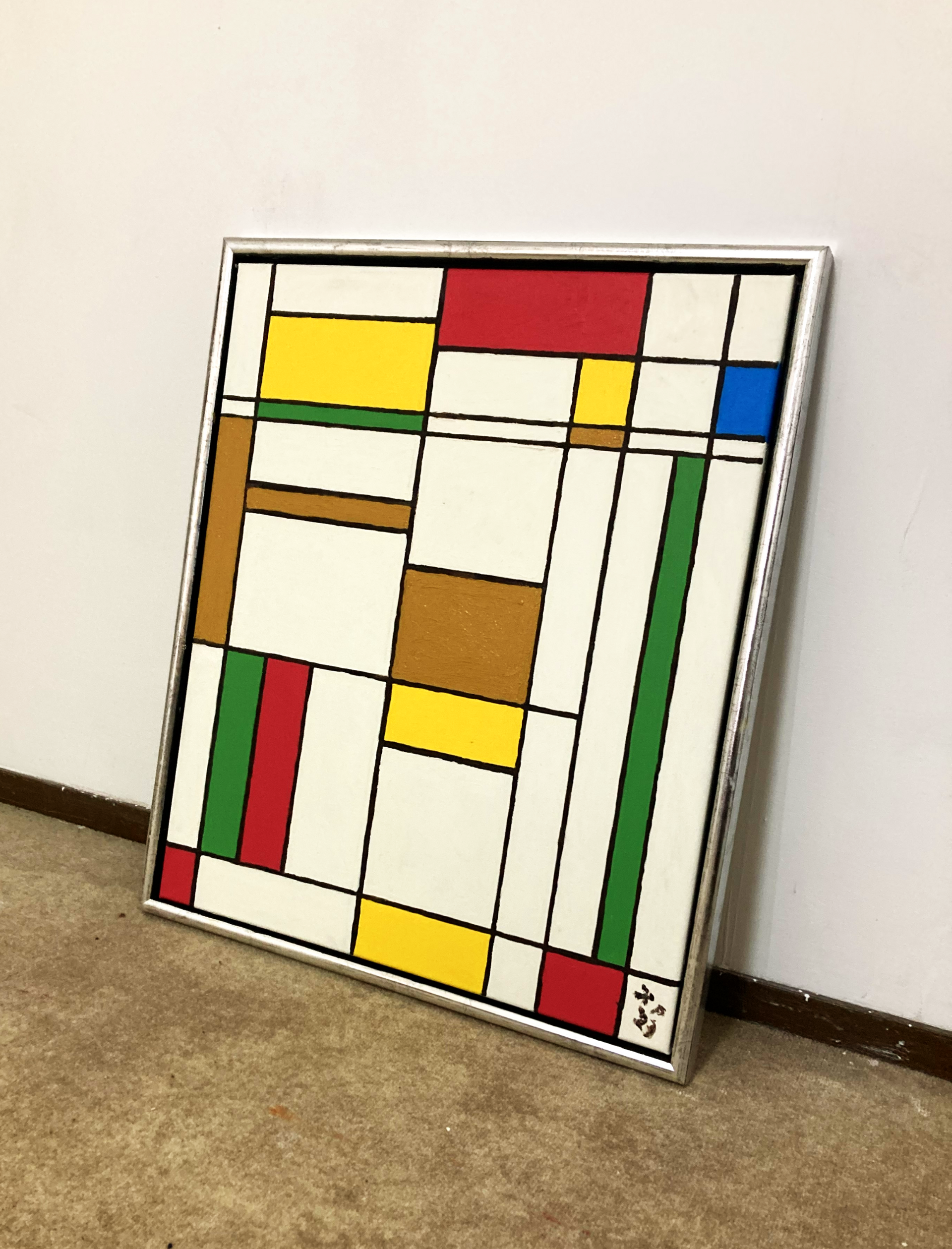 Abstract geometrisch schilderij in Mondriaan-stijl, in een zilveren lijst, leunend tegen een muur op een tapijt.