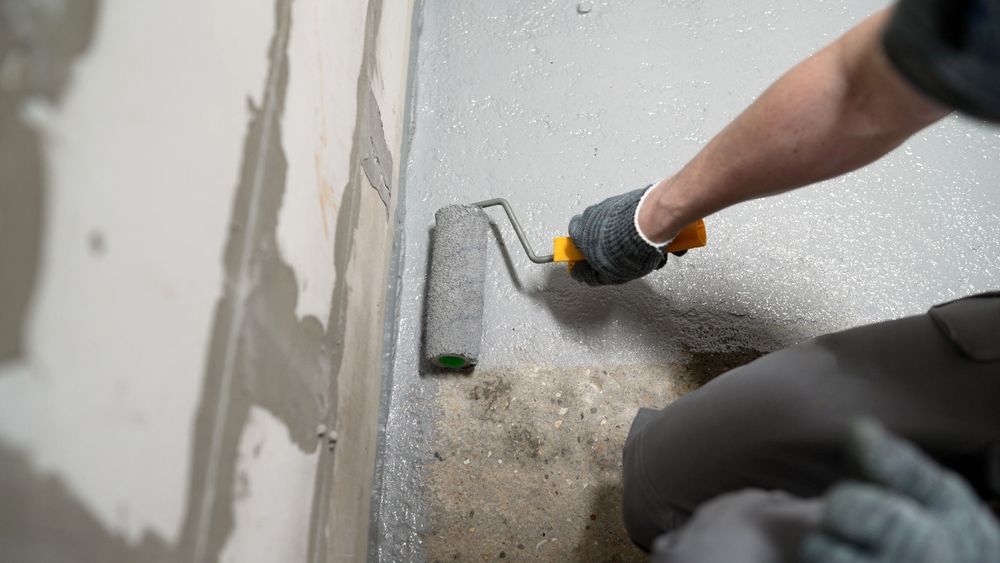 A person wearing work gloves uses a paint roller to apply gray coating to a concrete floor against a wall. — Hobart Tiling Solutions In Clarendon Vale, TAS