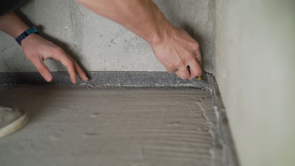 A person uses a utility knife to trim edge insulation tape along the wall of a room with a newly applied screed floor. — Hobart Tiling Solutions In Clarendon Vale, TAS