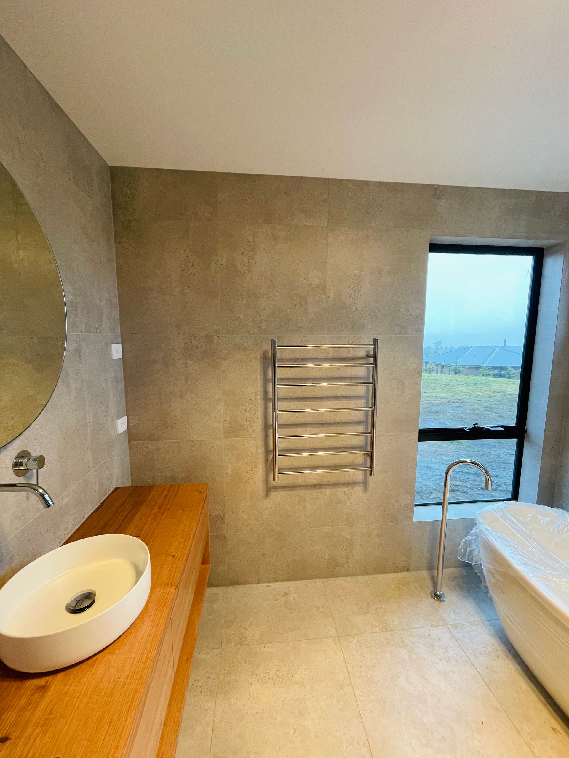 A modern bathroom with a stone-top vanity, vessel sink, wall-mounted towel warmer, and a freestanding tub by a window. — Hobart Tiling Solutions In Clarendon Vale, TAS
