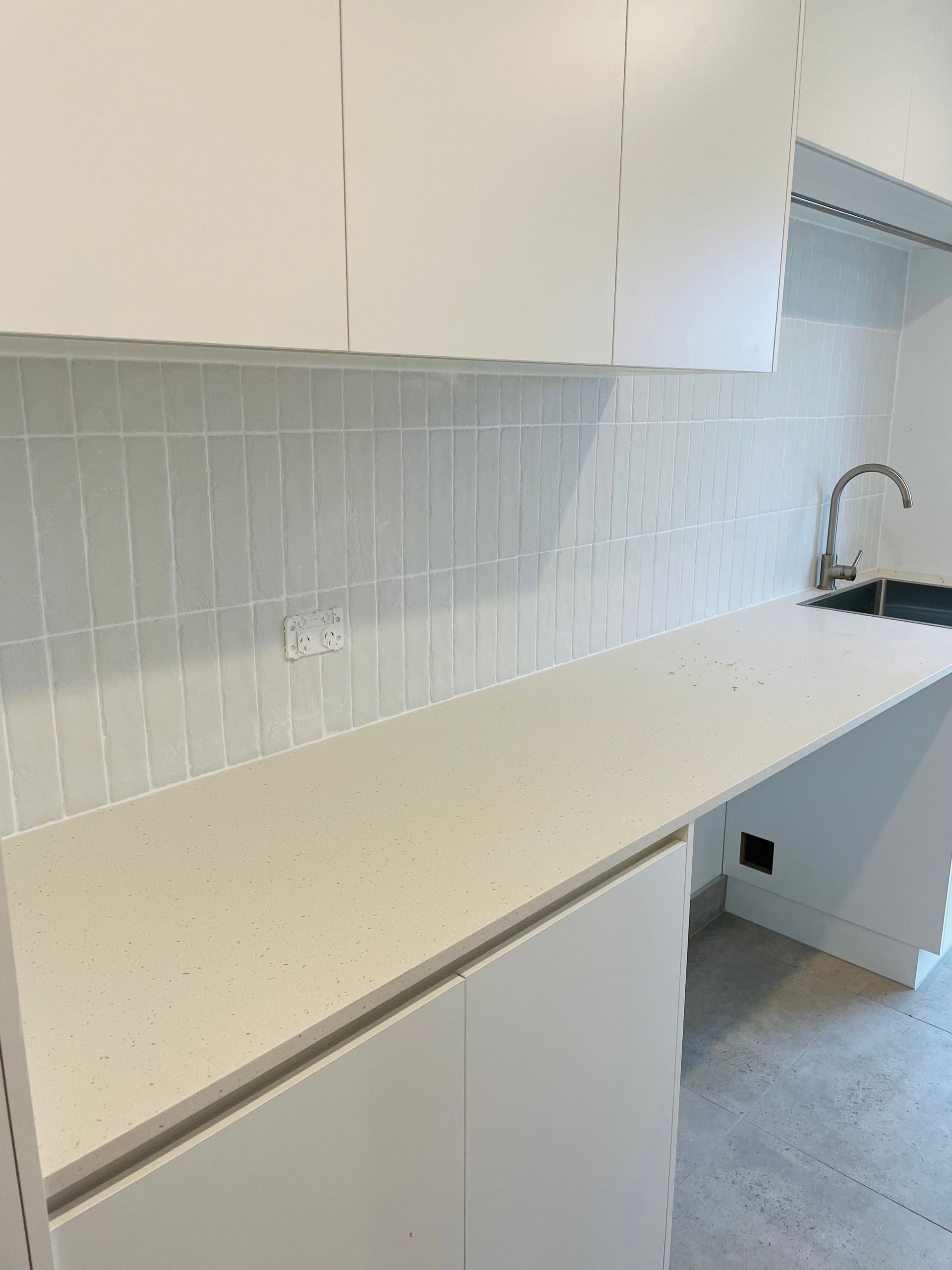 A modern, minimalist kitchen featuring white cabinets, a light-speckled countertop, and vertical white tiled backsplash — Hobart Tiling Solutions In Clarendon Vale, TAS
