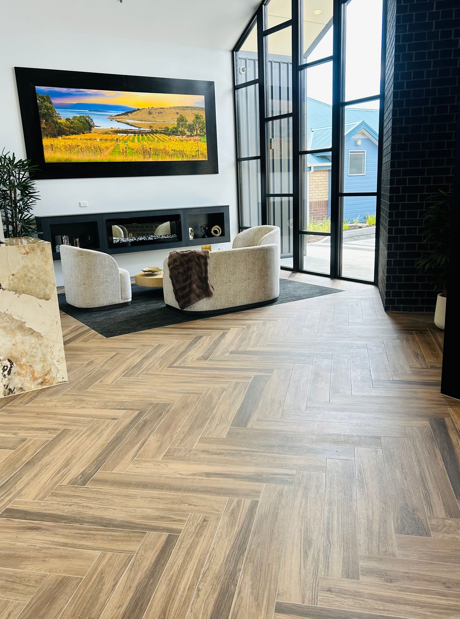 A modern living room with herringbone wood floors, two beige armchairs, a fireplace, and a large landscape painting. — Hobart Tiling Solutions In Clarendon Vale, TAS