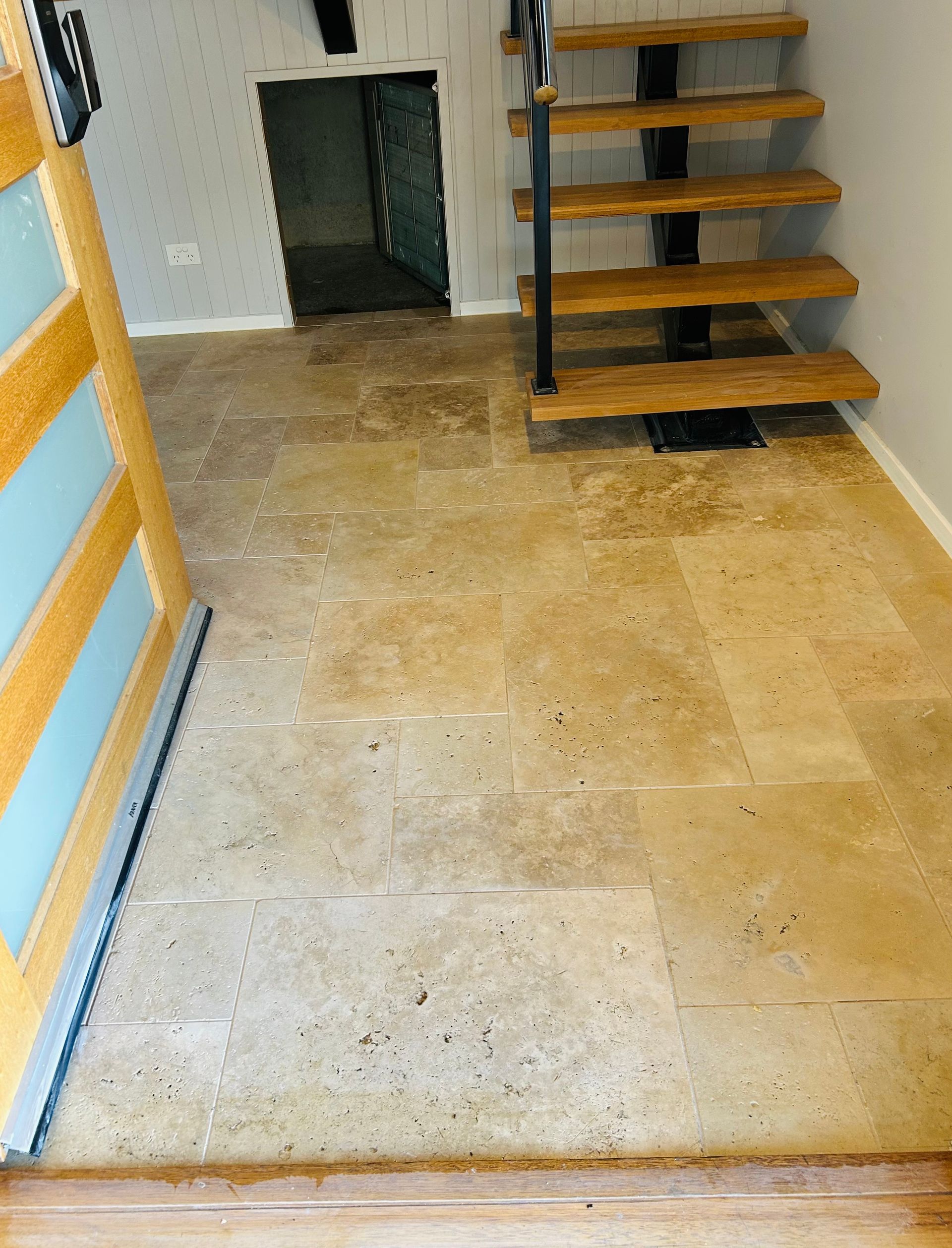 Tan stone tile flooring leading to a small doorway and a floating wooden staircase with a black metal railing. — Hobart Tiling Solutions In Clarendon Vale, TAS