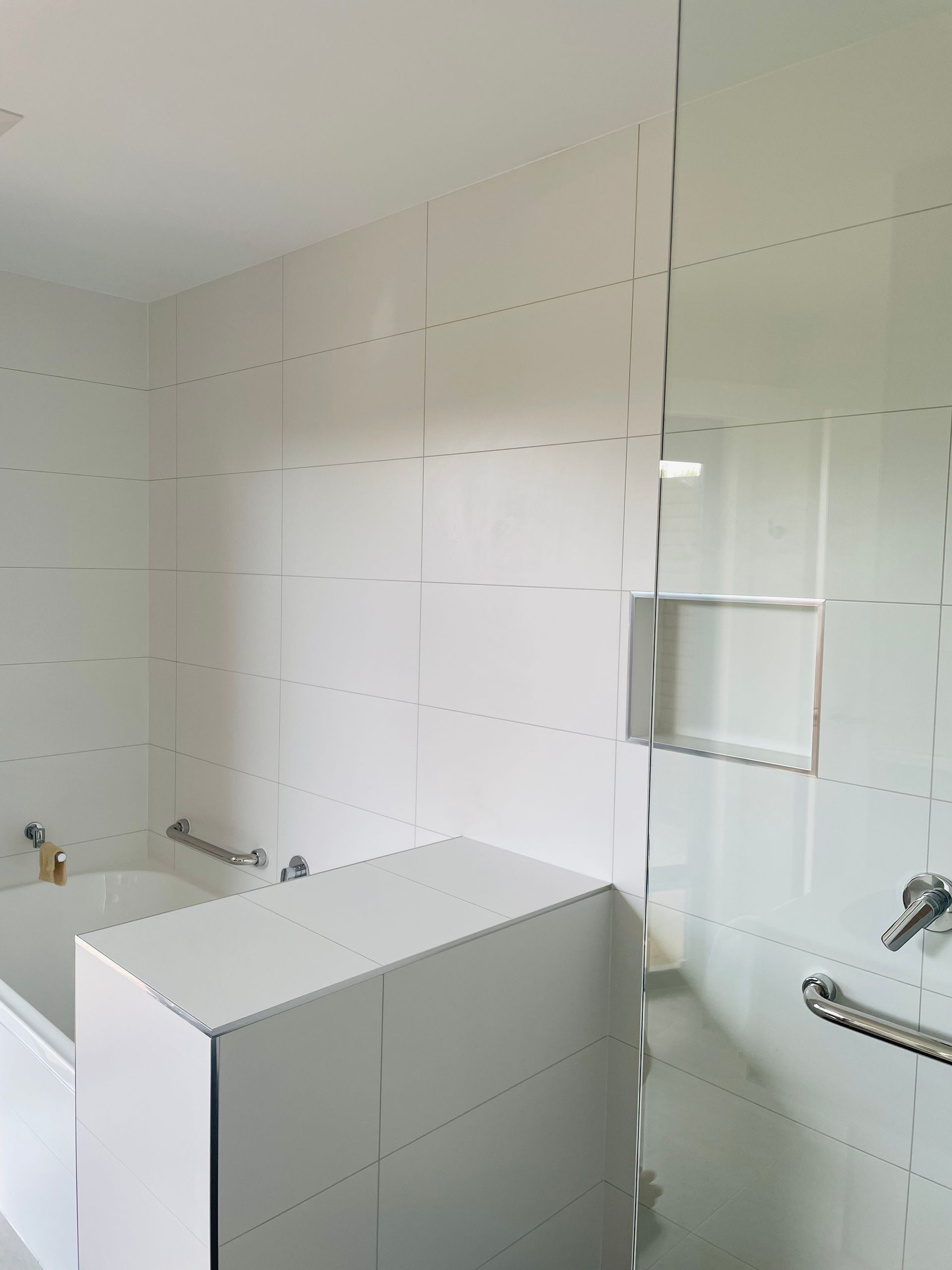 A bright, modern bathroom featuring white tiled walls, a built-in shelf, a glass partition, and safety grab bars. — Hobart Tiling Solutions In Clarendon Vale, TAS
