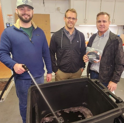 Three men are standing around a trash can and one of them is holding a hose