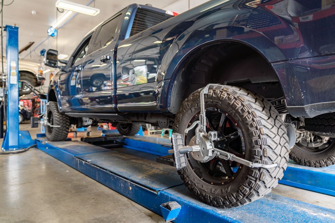 Blue pickup truck lifted on a car hoist in an auto repair shop, with off-road tires.