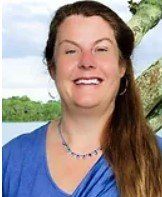 A woman wearing a blue shirt and a necklace is smiling in front of a tree.