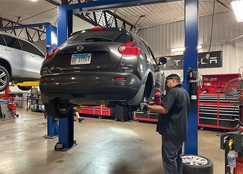 Black SUV lifted on a car hoist in an auto repair shop, with a mechanic working nearby.