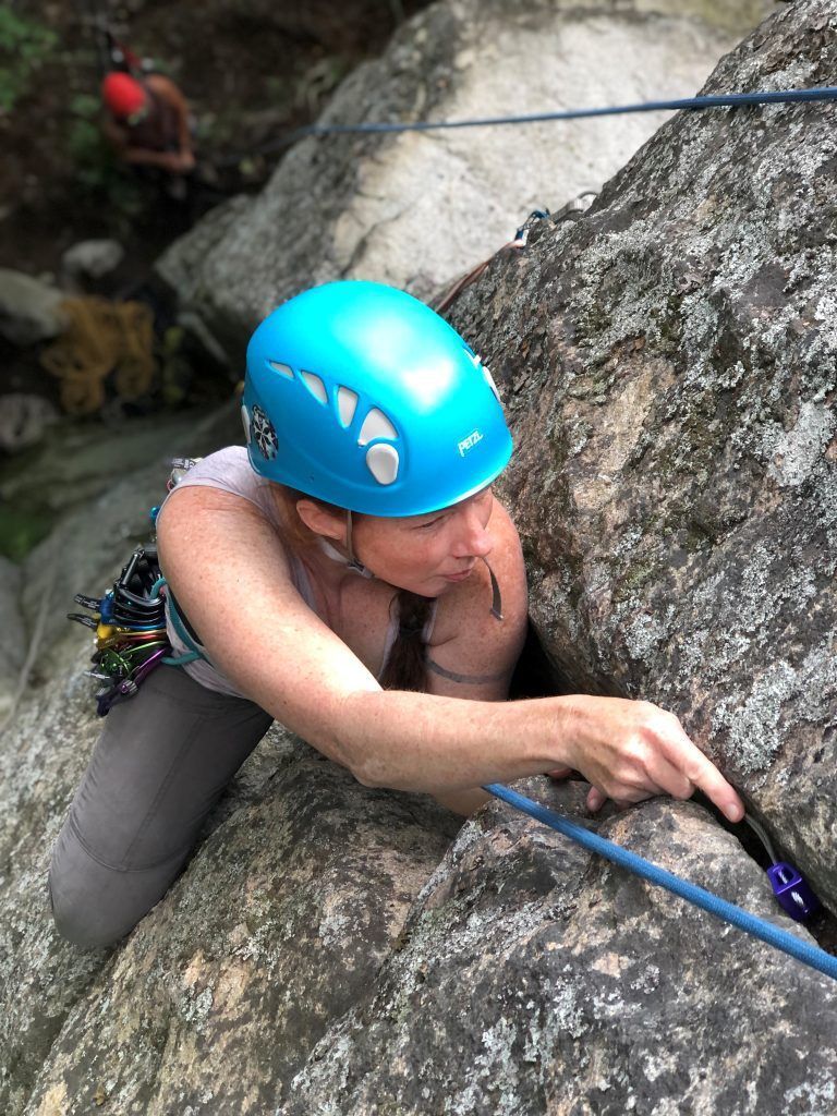 Woman rock climbing, wearing a blue helmet and harness, reaching for a handhold on a gray rock face.