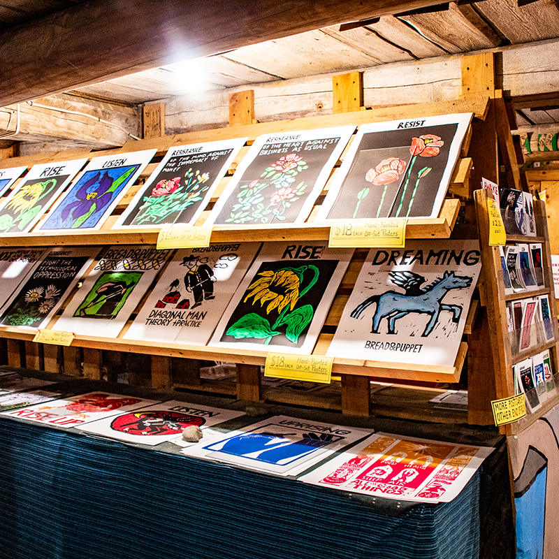 Posters on display at a Bread and Puppet VT; wooden shelves, various colors, floral and graphic designs.
