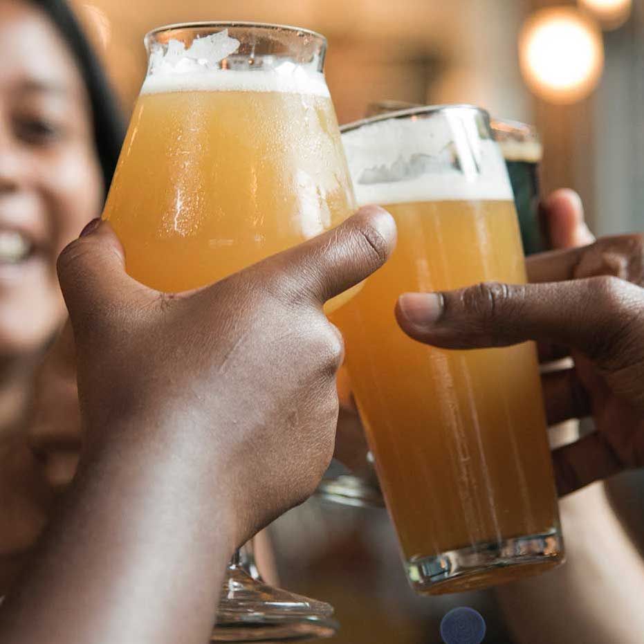 People toasting with beers, close-up of hands holding glasses with amber and dark brews; blurred background.