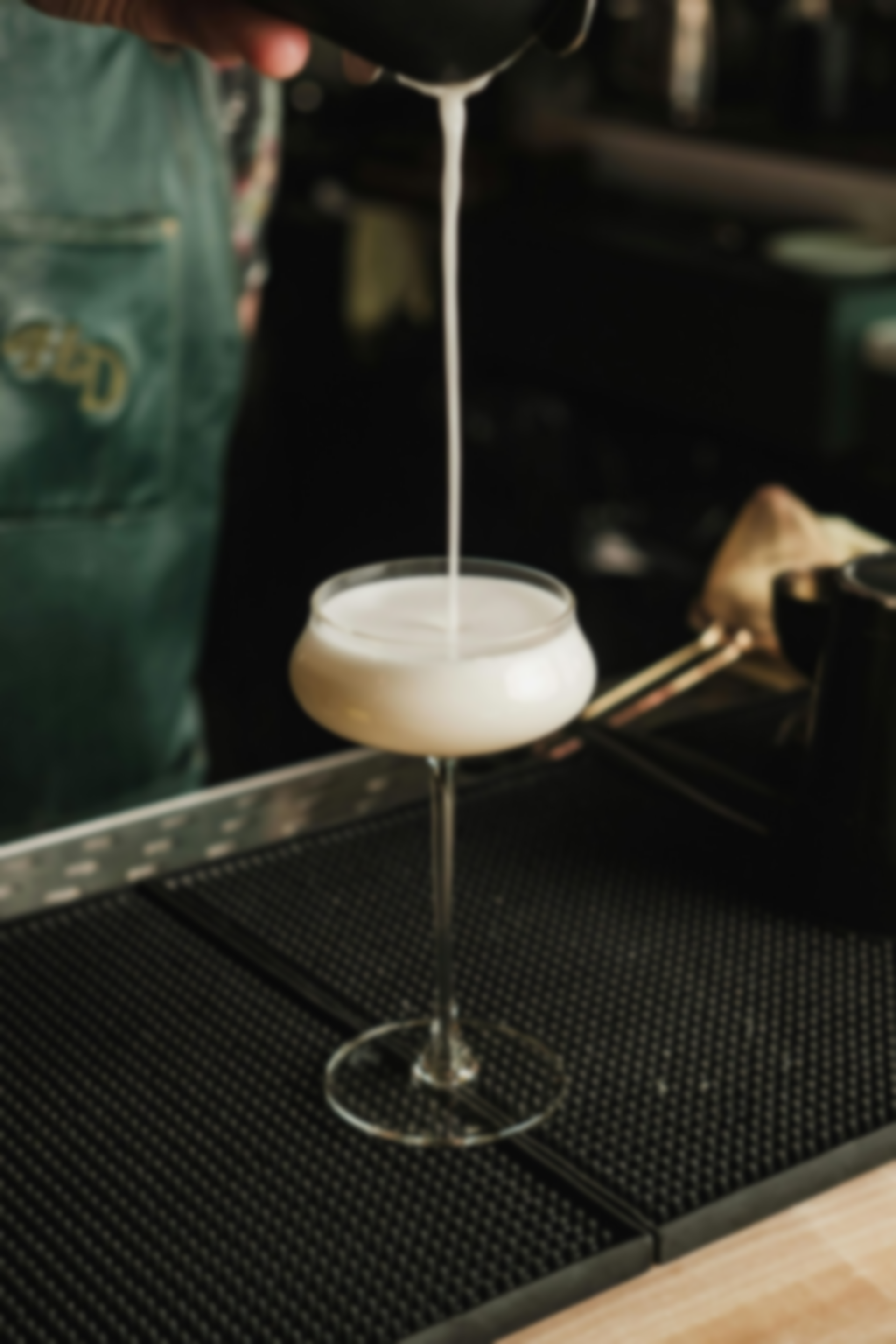 A bartender pouring a white cocktail into a stemmed glass. The drink is creamy with a dark bar setting.