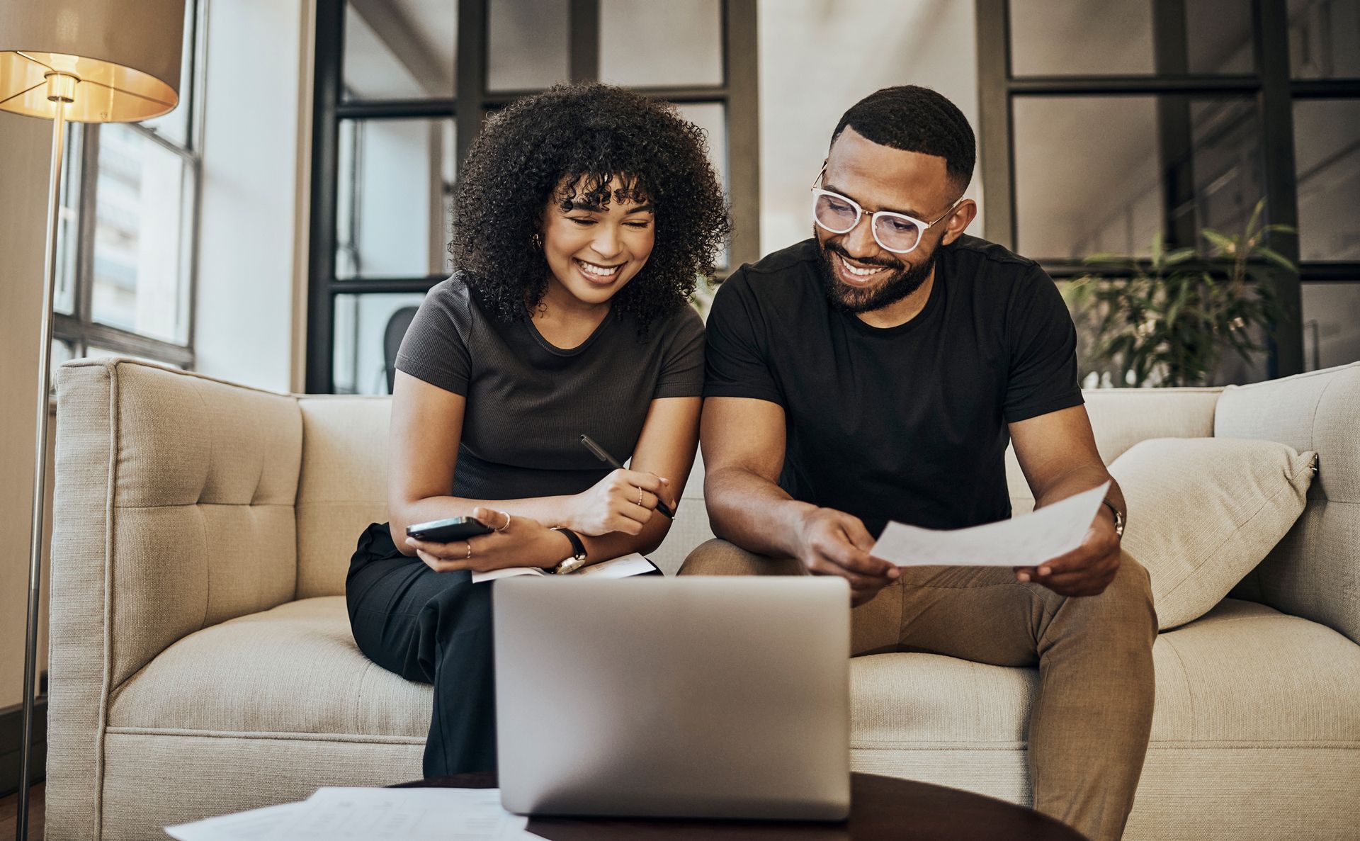 A smiling couple is seated on their coach, reviewing in their laptop their personal loan.