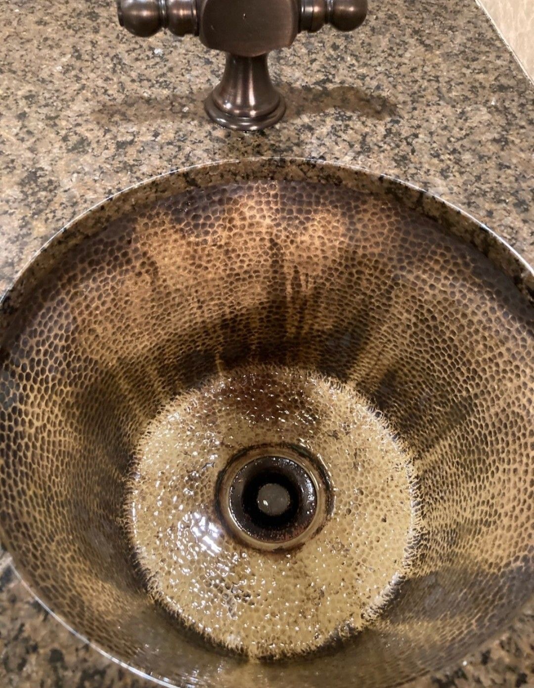 Close-up of a round, textured glass vessel sink with a drain, set in a granite countertop.