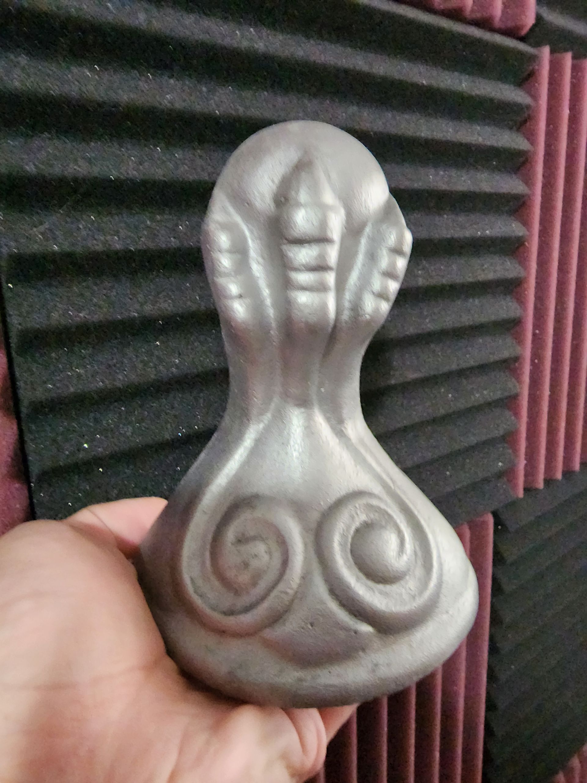 Hand holding a small silver figurine with spiral designs, in front of black soundproof foam panels.