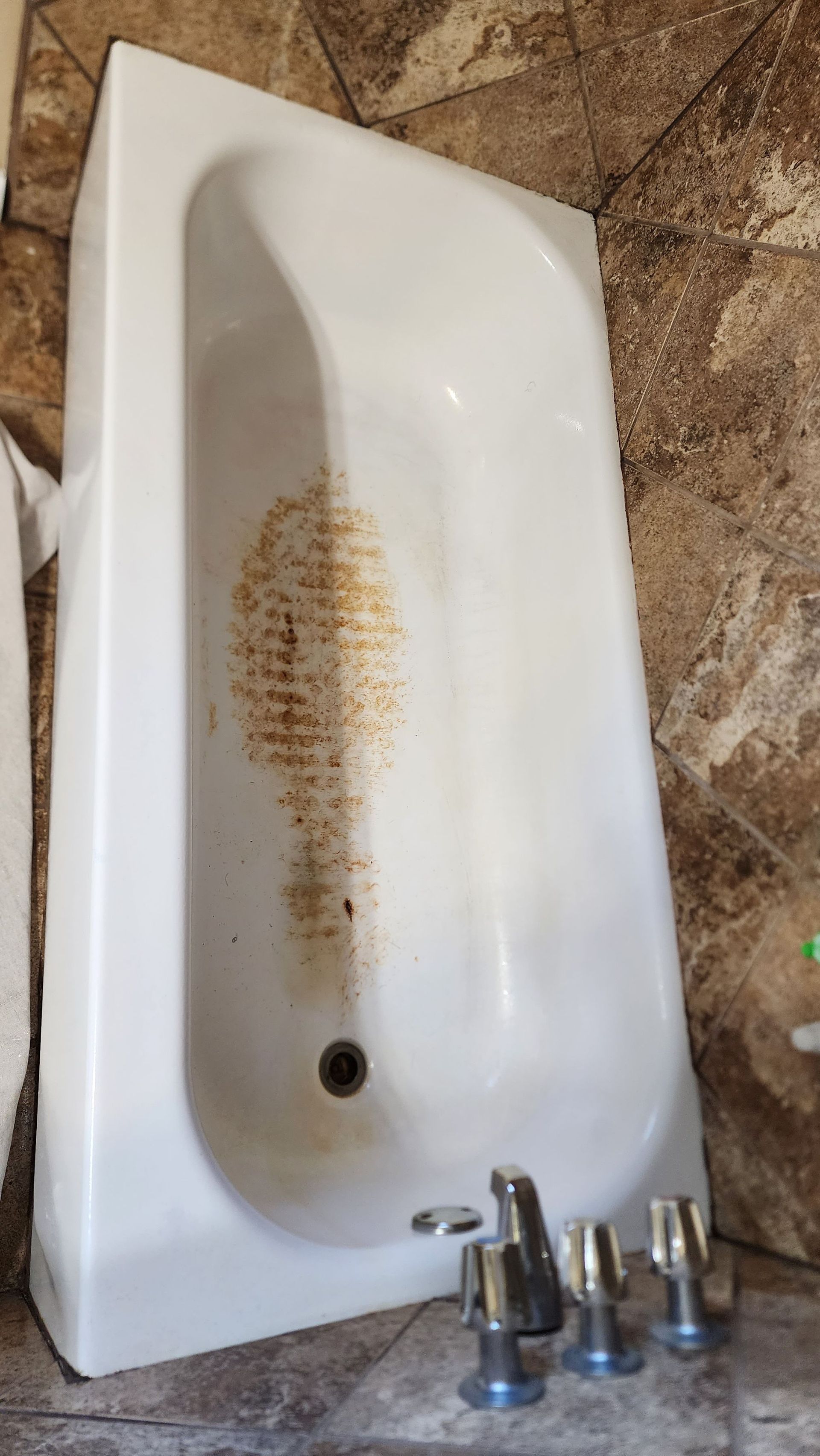 Dirty white bathtub with brown stains and three metal faucet handles on the floor nearby