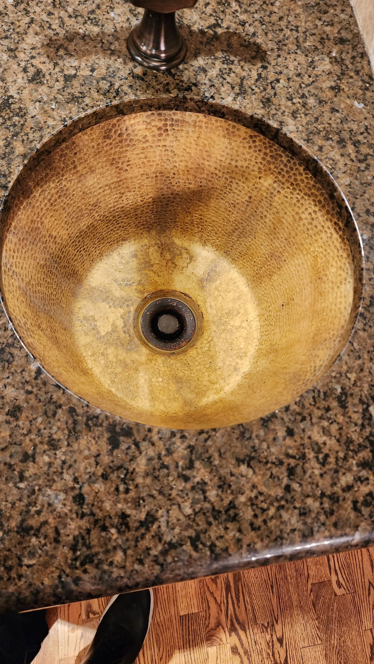 Round beige stone sink with a dark drain, set in a speckled granite countertop.