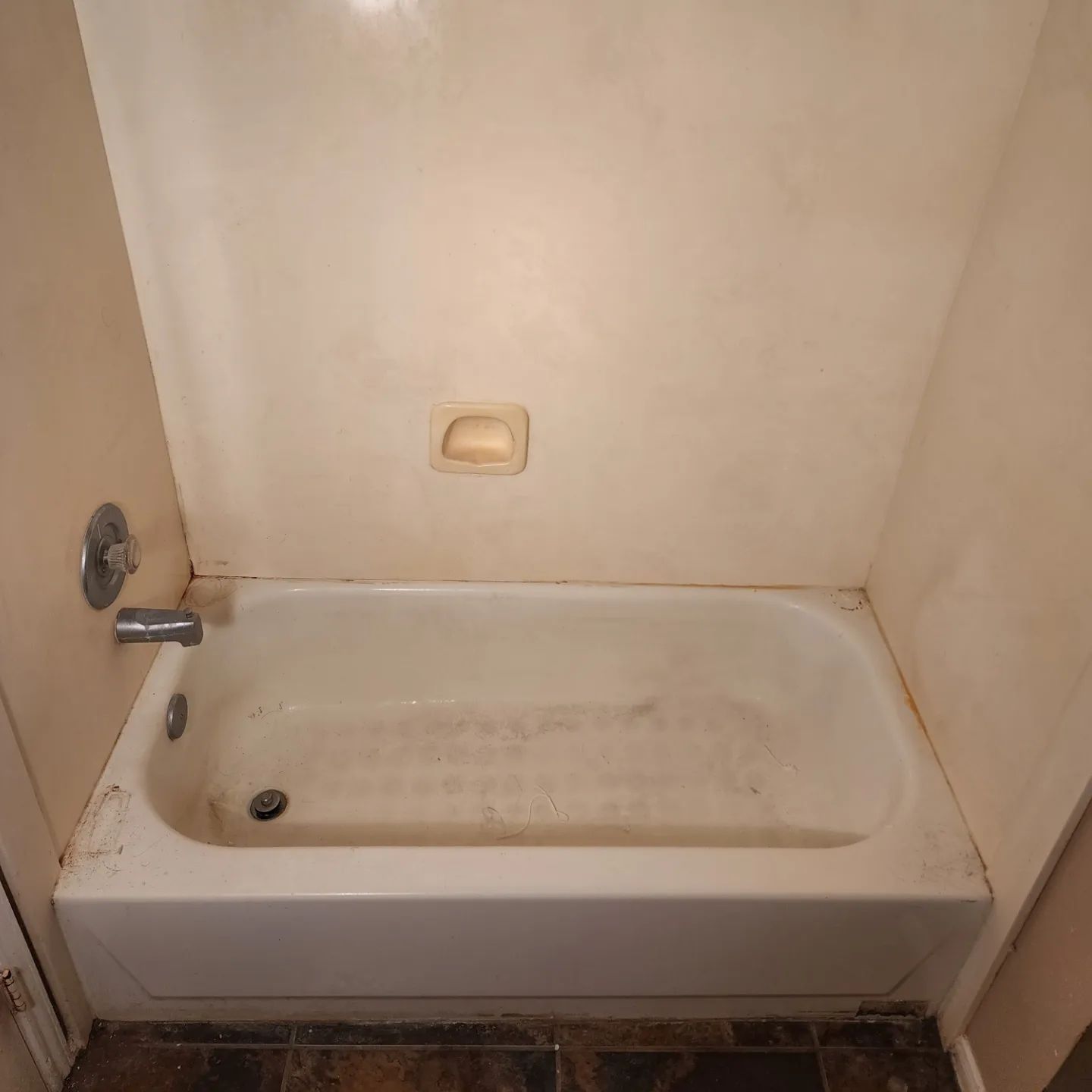Small beige bathtub with shower handle and grab bar in a plain bathroom.
