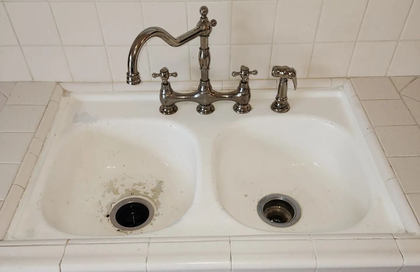 White double sink with chrome faucets and dirty drains in a tiled bathroom.