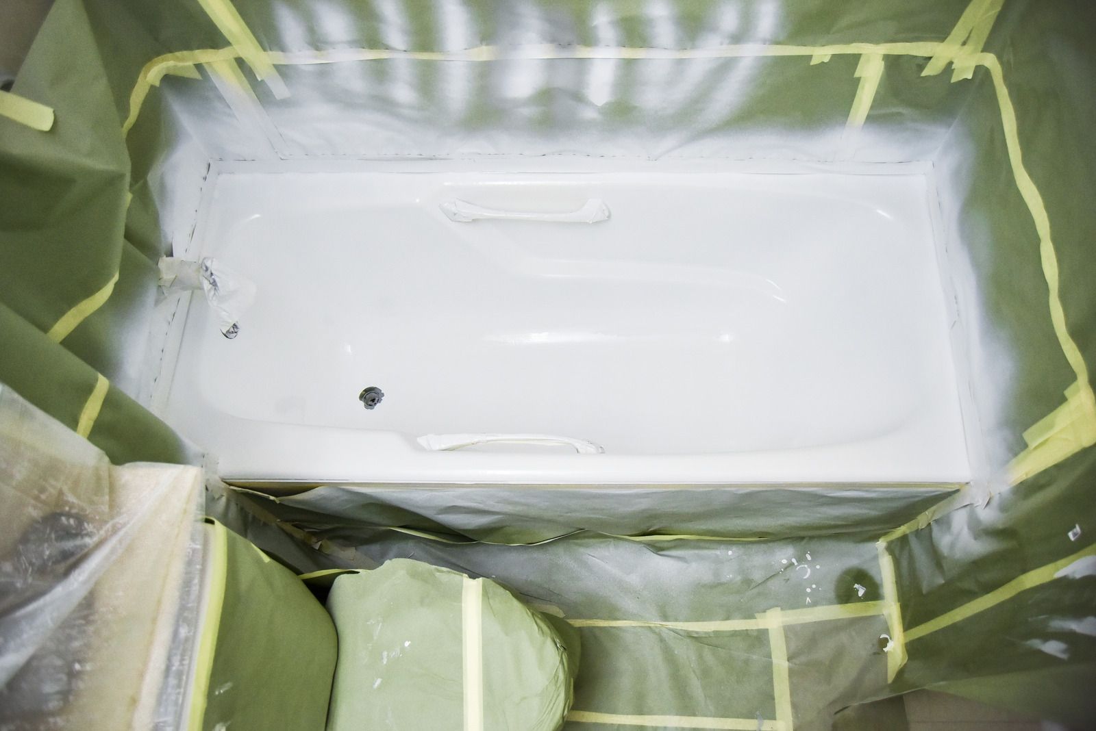 White bathtub being painted or refinished, masked with green tape and plastic sheeting.