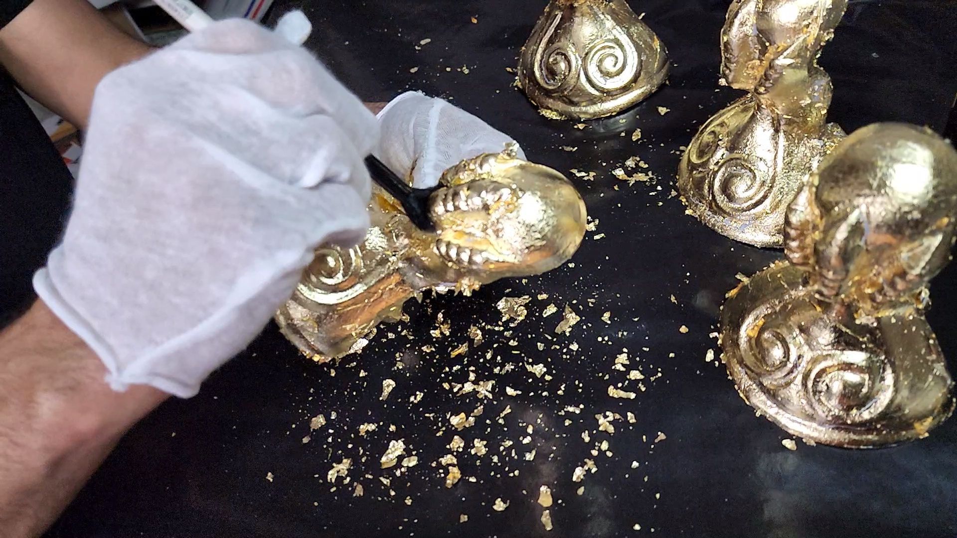 Gloved hand cleaning ornate gold figurines on a black surface with scattered dust