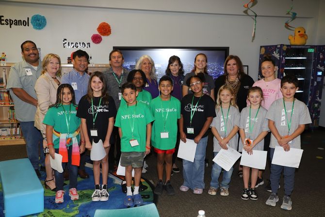 Group of children and adults posing in a classroom, several children holding papers and green shirts.