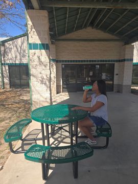 Person seated at a green picnic table outside a building, drinking from a cup.