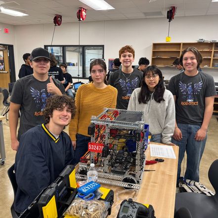 Six students pose beside a robotics competition robot in a classroom, with tools and parts on a table.