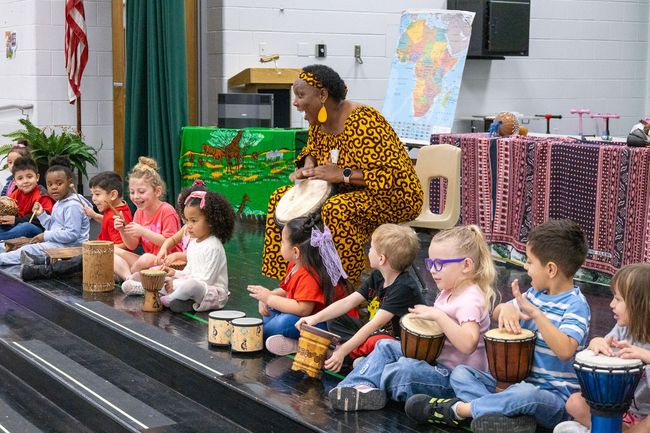 Children seated on a stage play drums while a teacher leads a music activity.