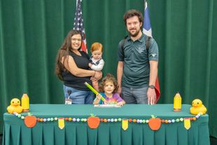 Family posing at a decorated table with a child and baby ducks, in front of green curtains and flags