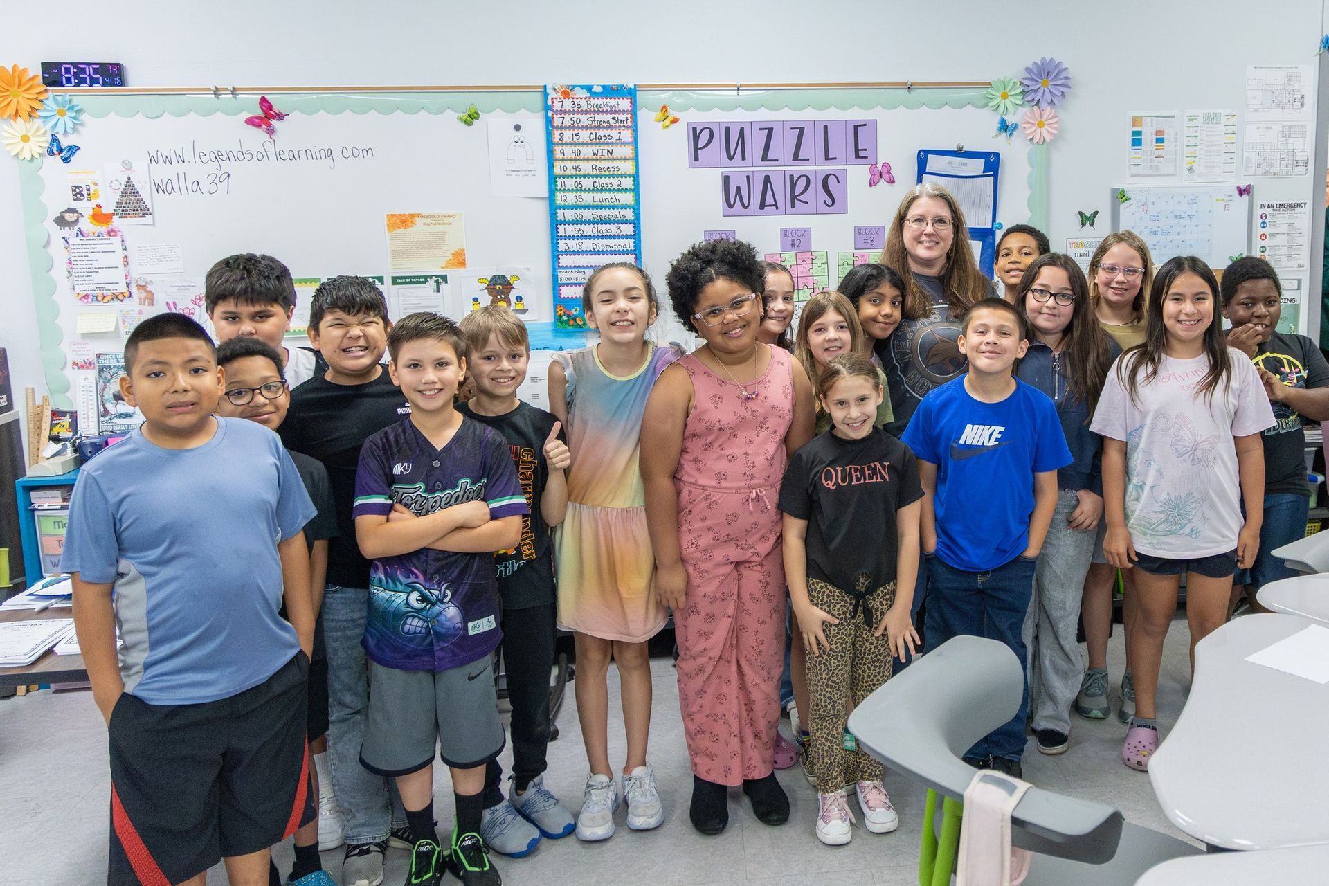 Group of smiling children and a teacher standing in a classroom beside a “Puzzle Wars” bulletin board.
