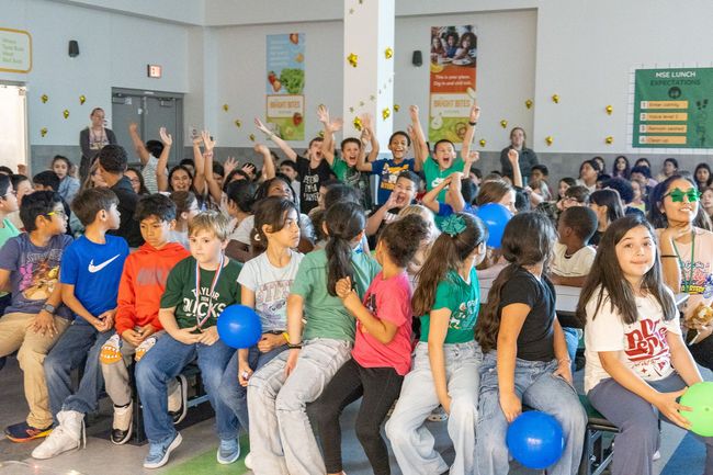 Students seated in rows, cheering and holding balloons in a bright indoor school hall