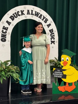 Child in teal graduation cap and gown stands beside an adult in a light green dress at a school photo backdrop.