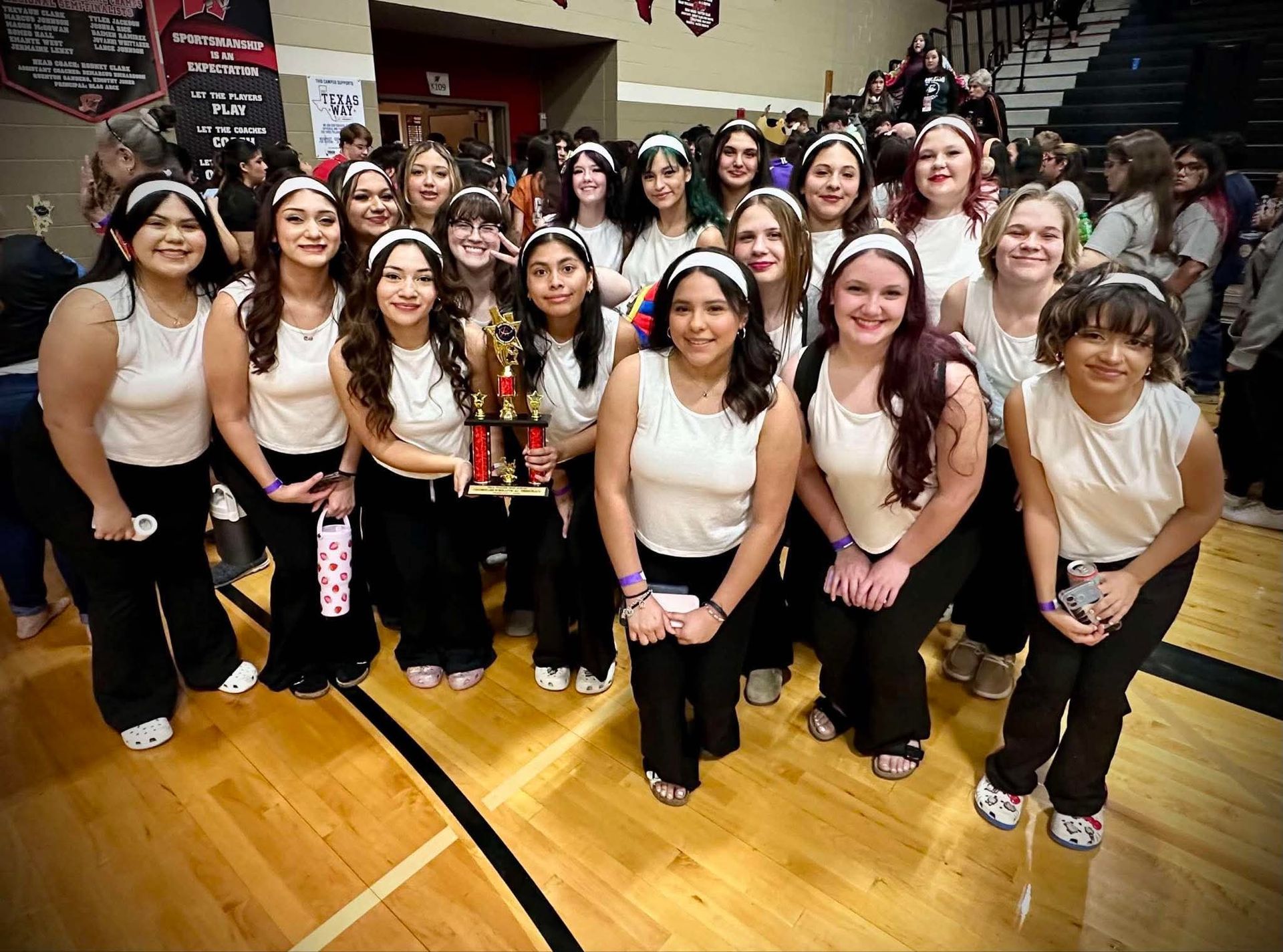 Group of students posing in a gym, wearing white tops and black pants, with smiles and bleachers behind them