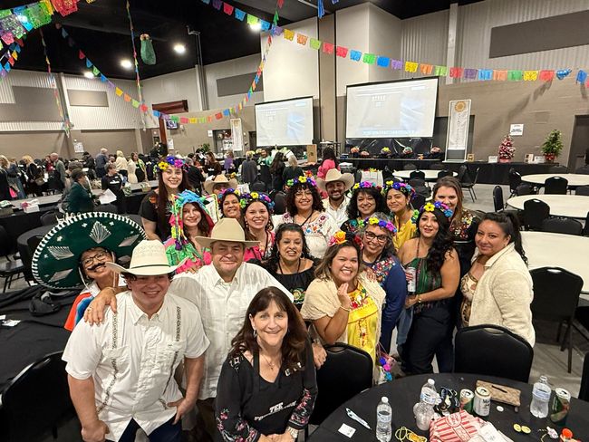 Large indoor group photo at a festive banquet, with smiling people at round tables under colorful pennants