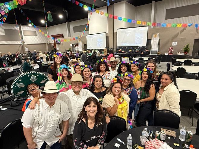 Large indoor group photo at a festive banquet, with smiling people at round tables under colorful pennants