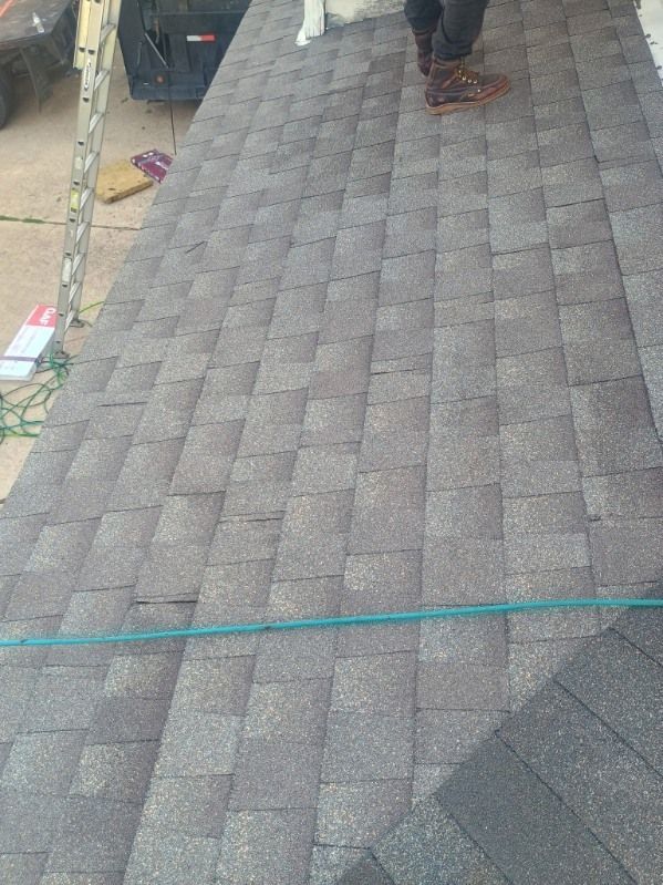 Roof shingles with a worker’s legs visible near the top and a green rope across the lower edge.