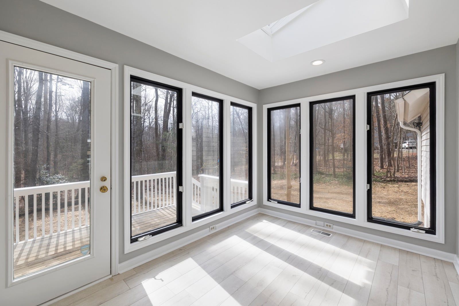 Bright sunroom with white walls, large corner windows, and a glass door overlooking a wooded deck.