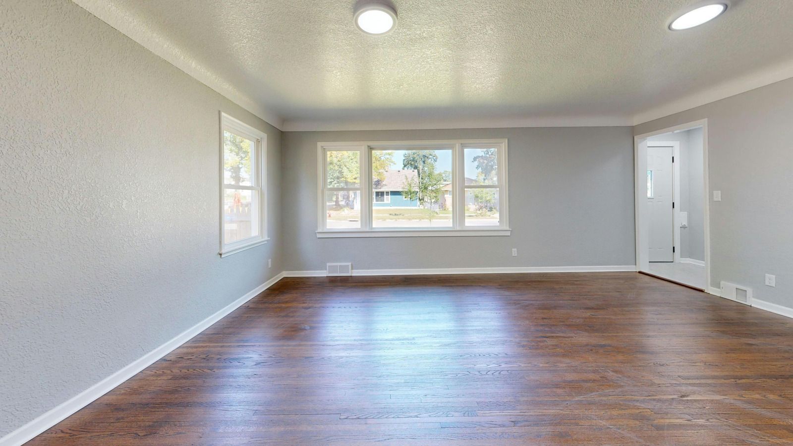 Empty living room with hardwood floors, white walls, recessed lights, and large front windows.