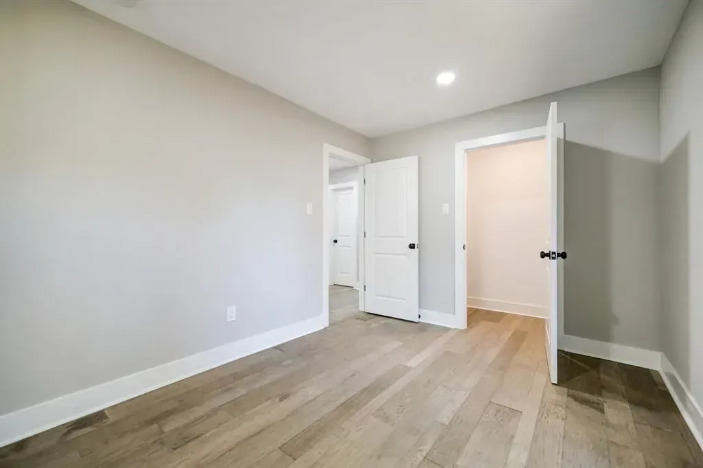 Empty bedroom with light gray walls, wood floors, and open white doors leading to a bathroom and hallway