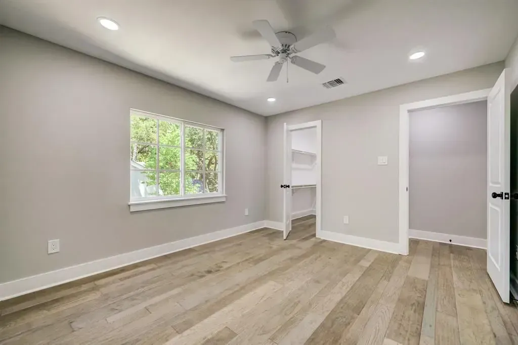Empty beige bedroom with wood floors, white trim, ceiling fan, and a window letting in daylight